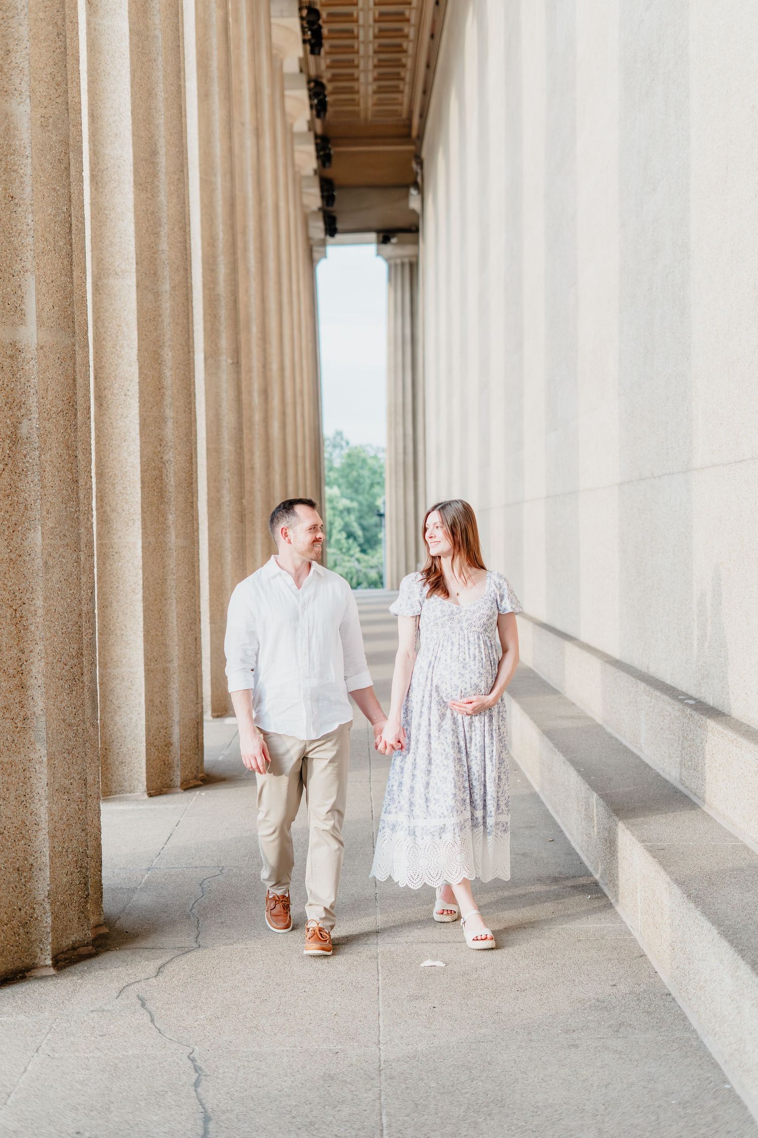A couple poses together near blooming hydrangeas with a classical stone building visible among lush green trees behind them.