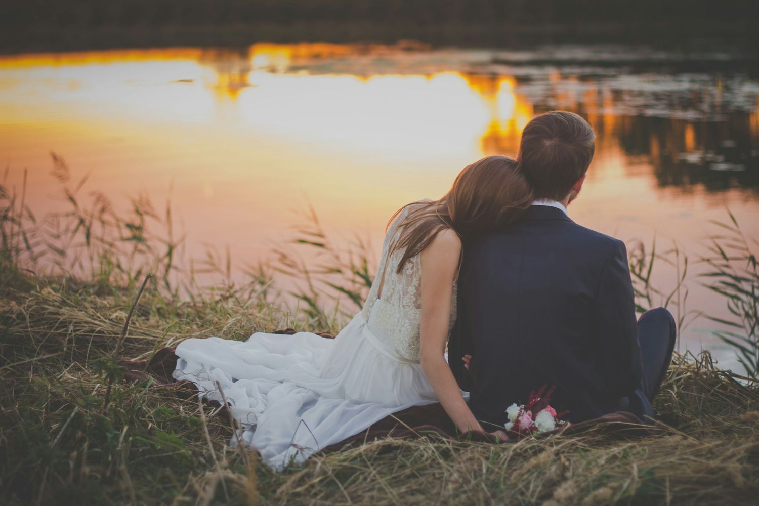 Newlywed couple sitting together by a serene lake at sunset, bride in white dress with bouquet, groom in navy suit.