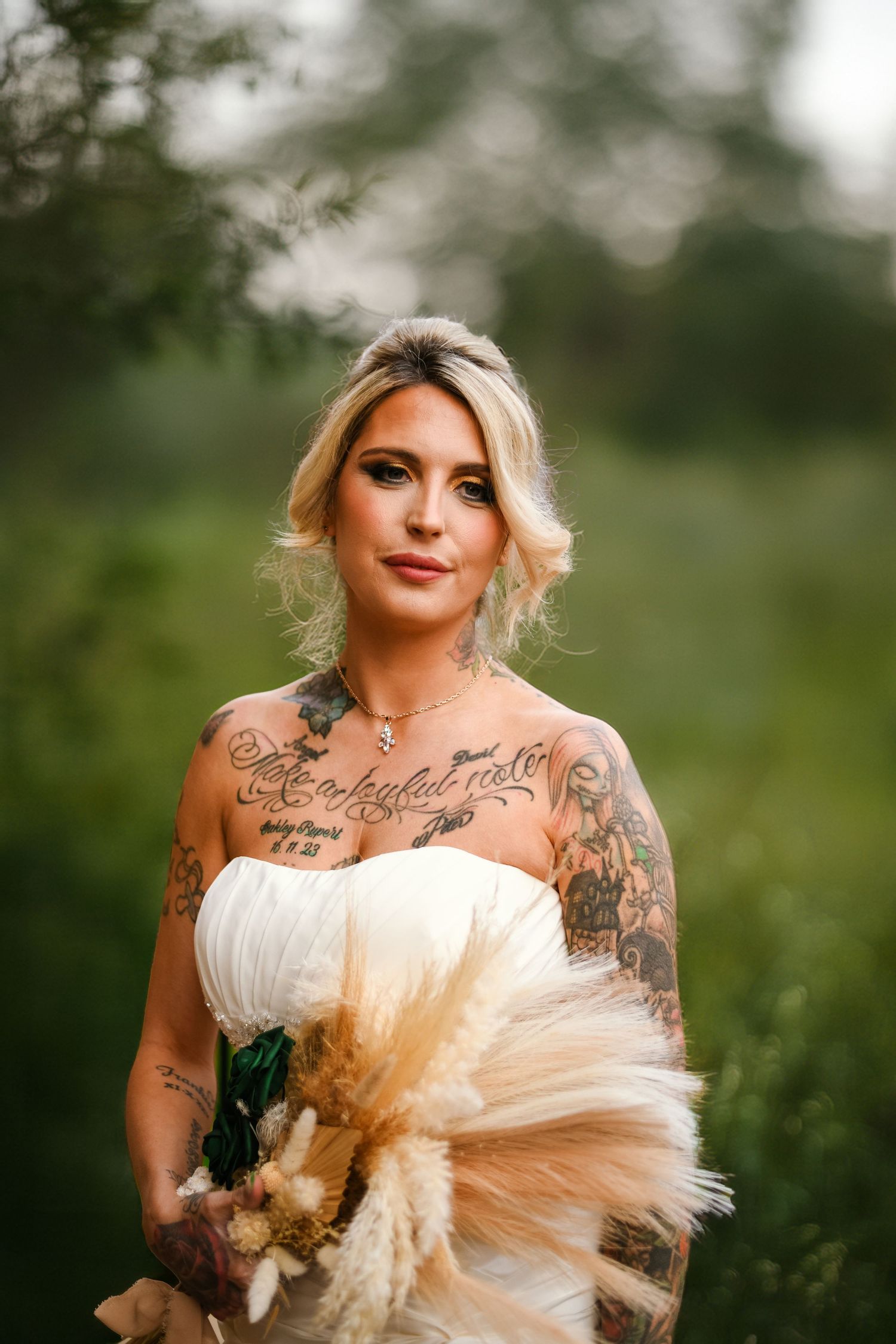 Tattooed bride in strapless white gown holding pampas grass bouquet, posing outdoors in lush green nature setting.
