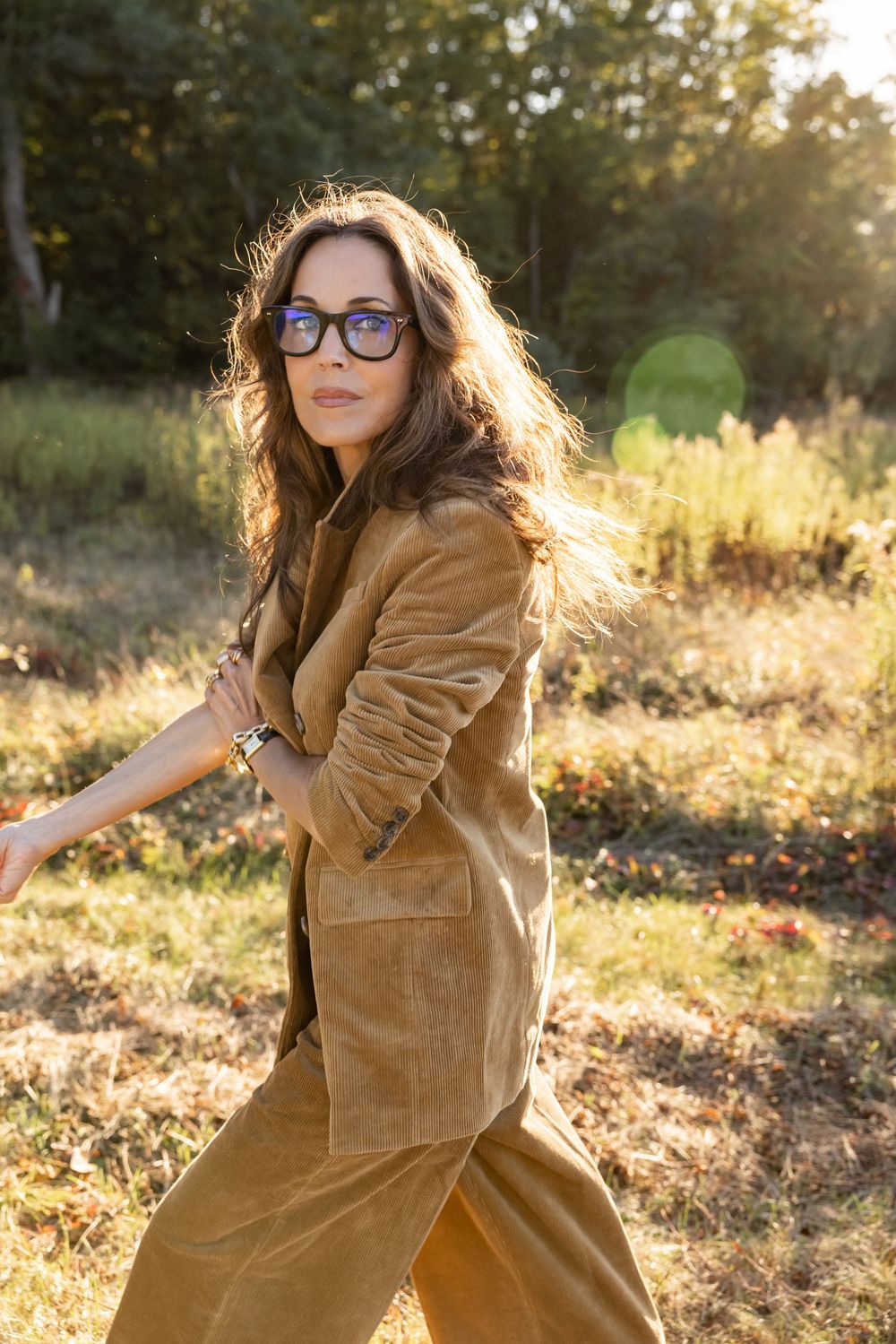 Woman in tan corduroy suit and sunglasses walks through sunlit outdoor field.