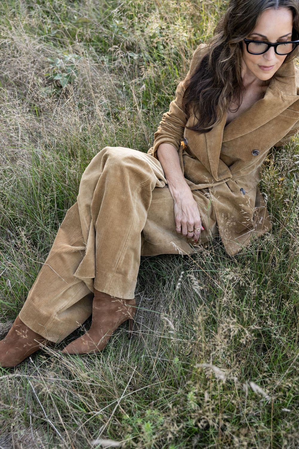 Woman in tan corduroy suit and brown boots sits relaxed in tall grass outdoors.