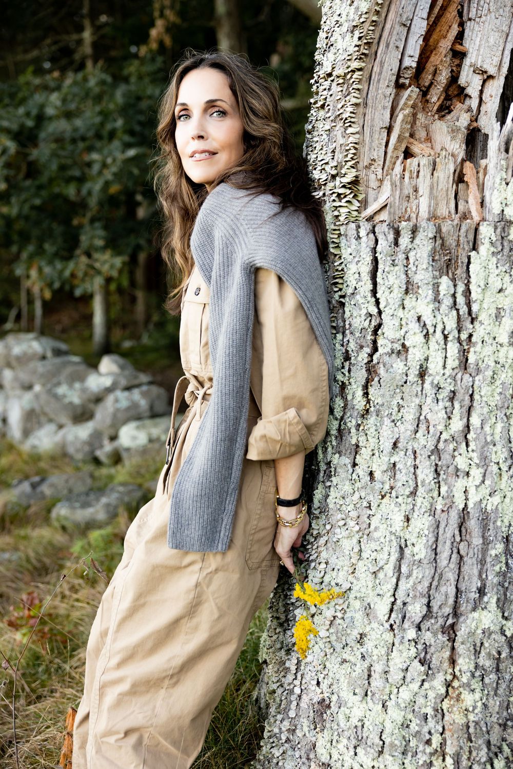 Woman in beige belted jumpsuit with gray scarf leans against a lichen-covered tree.