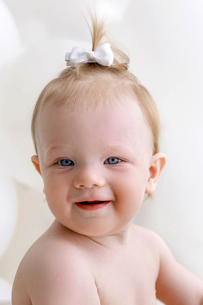 Smiling baby girl with blue eyes and white bow hair clip, portrait photo on light background.