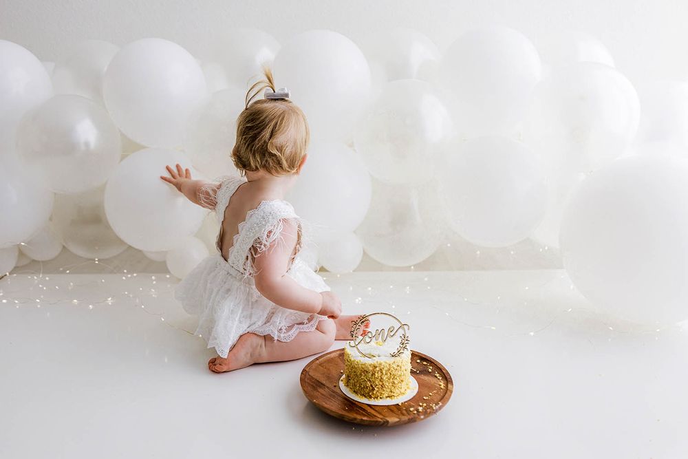 Baby girl in white lace dress reaching toward white balloons beside a smash cake on a wooden plate.