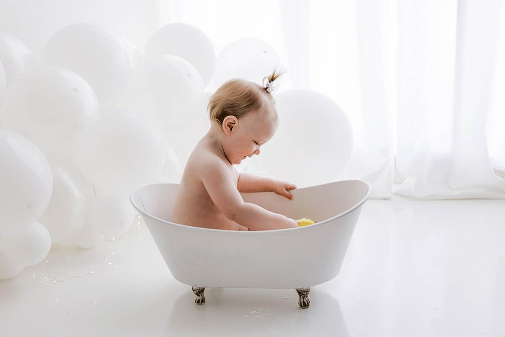 Baby sitting in a small white bathtub playing with a yellow toy, surrounded by white balloons in a bright studio.