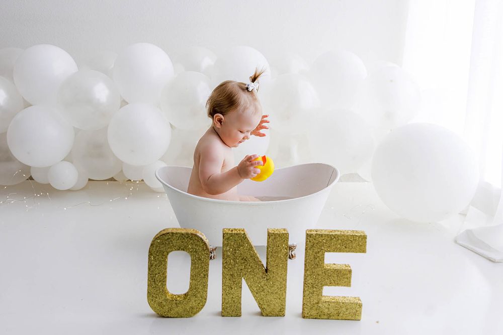 Baby girl sits in white tub with rubber duck, gold ONE letters and white balloons for first birthday photo.