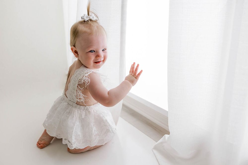 Smiling baby girl in white lace dress kneeling by bright window, waving hand in airy white room.