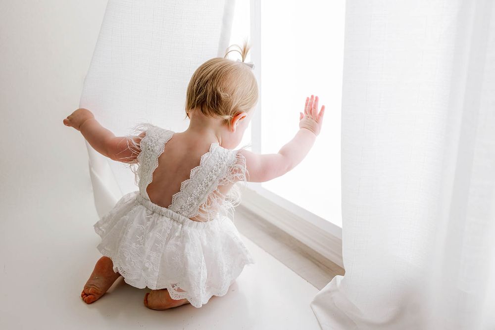 Baby girl in white lace romper kneeling by a bright window, reaching toward sheer curtains.