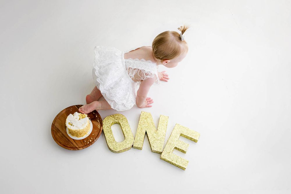 Baby girl in white dress touches first birthday cake next to gold glitter ONE letters on white background.