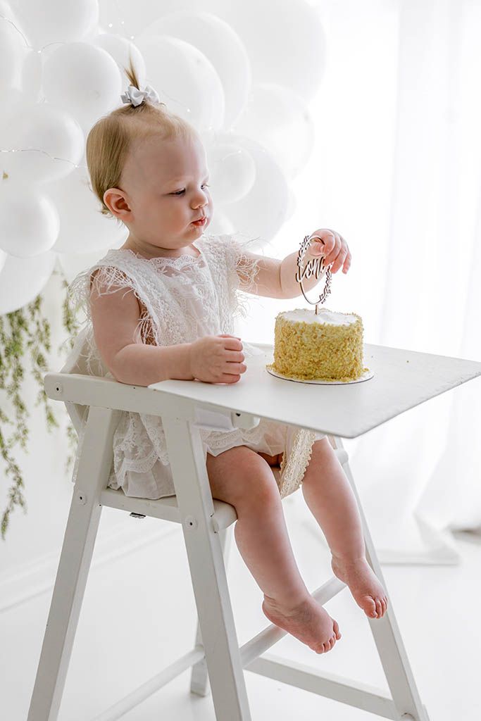 Baby girl in white lace outfit sits in high chair touching her first birthday cake surrounded by white balloons.