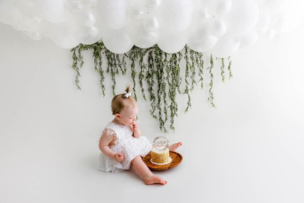 Baby girl in white lace dress sitting beside smash cake under white balloon and greenery backdrop.