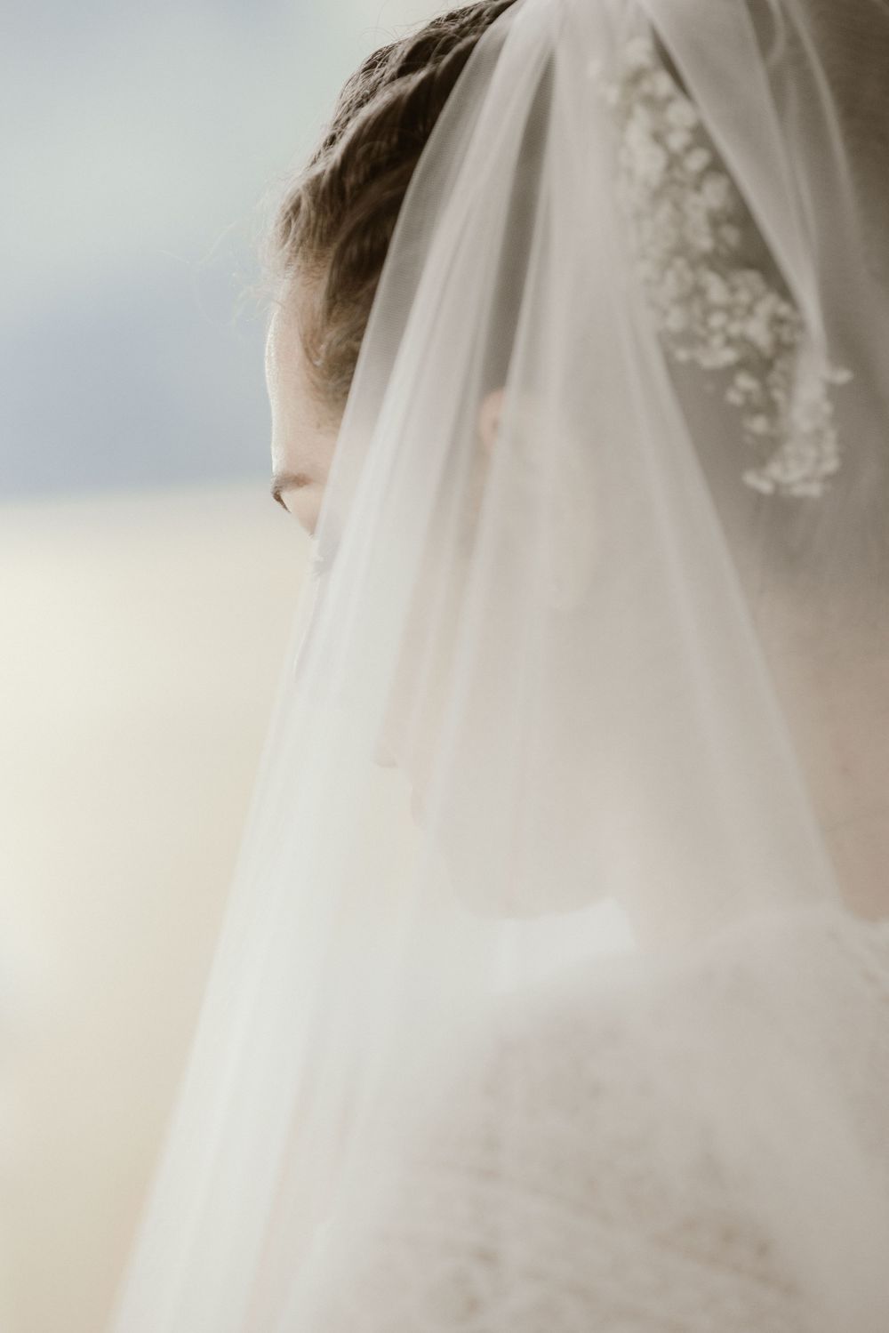 A bride wearing a delicate white veil adorned with small floral details, partially covering her face in a soft, dreamy portrait.