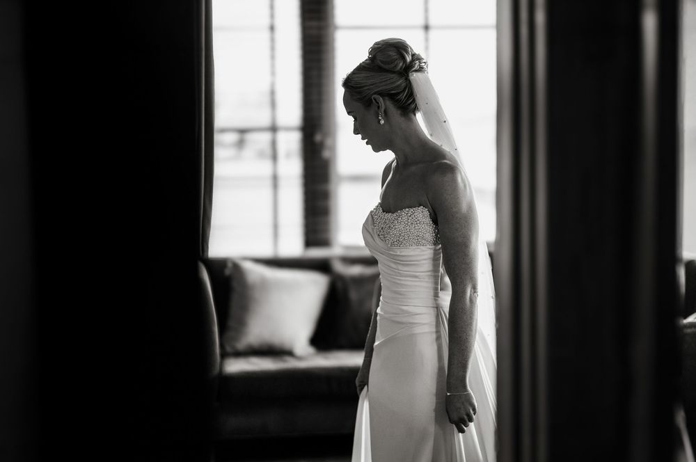 Black and white photo of a bride in a strapless wedding gown with veil, standing by a window in soft light.