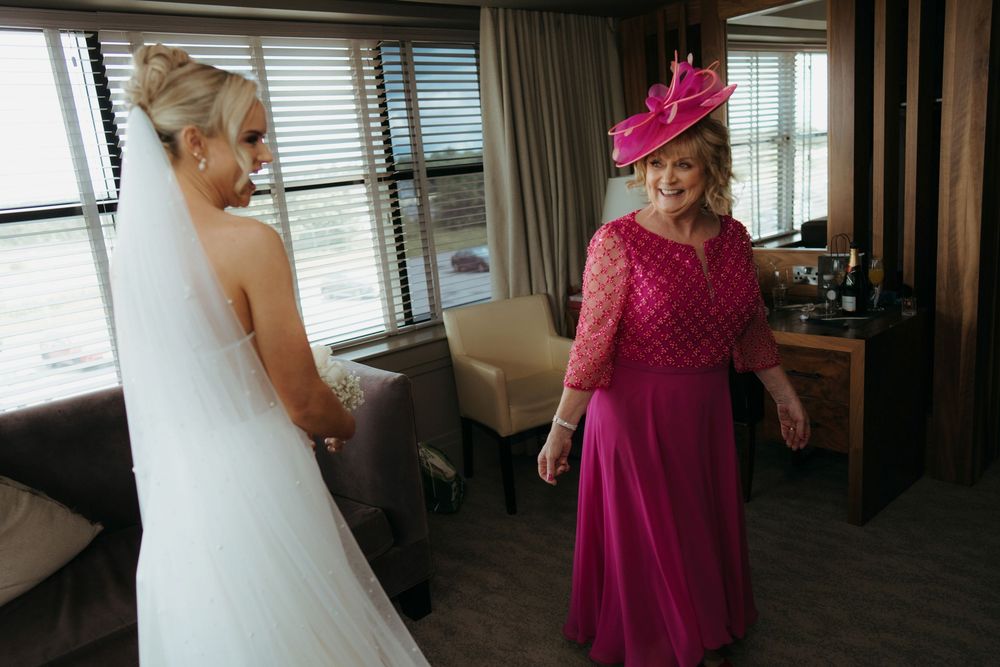 Bride in white gown admired by woman in pink dress and hat in hotel room wedding preparation.