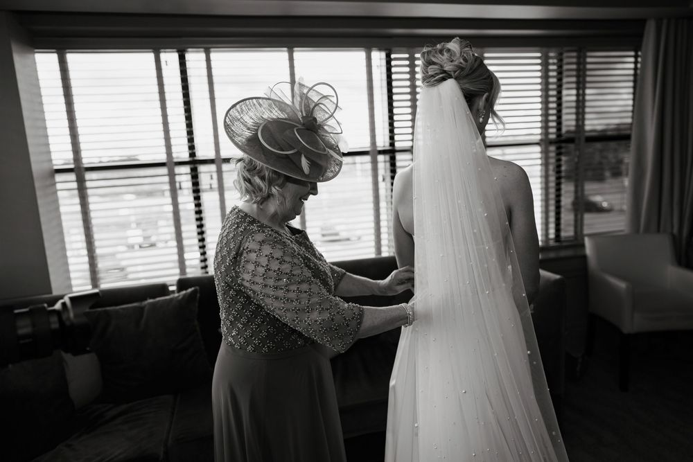 Black and white photo of a bride having her wedding dress fastened by an older woman in a hat near a window.
