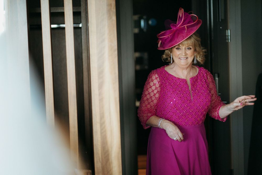 Woman in elegant fuchsia dress and matching fascinator hat poses gracefully in a doorway.