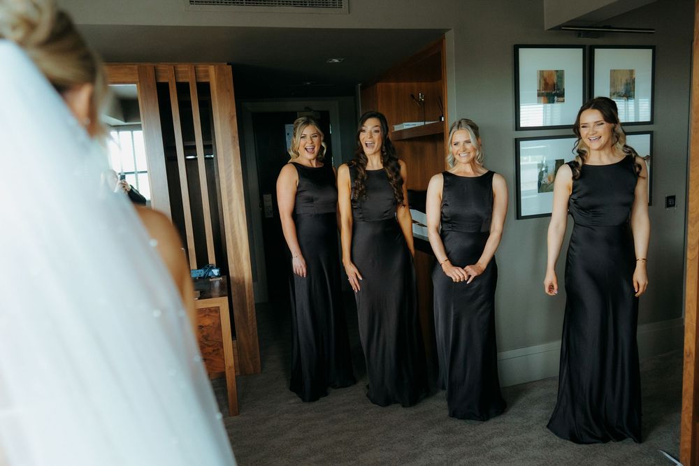 Bridesmaids in elegant black gowns react with joy as bride in white wedding dress and veil reveals her look in hotel room.
