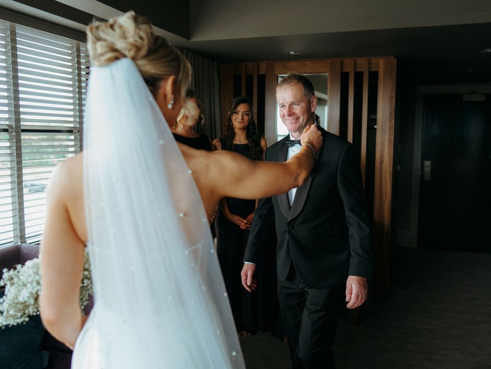 Bride in white veil reaches to adjust groom's boutonniere as bridesmaids look on during wedding day first look.