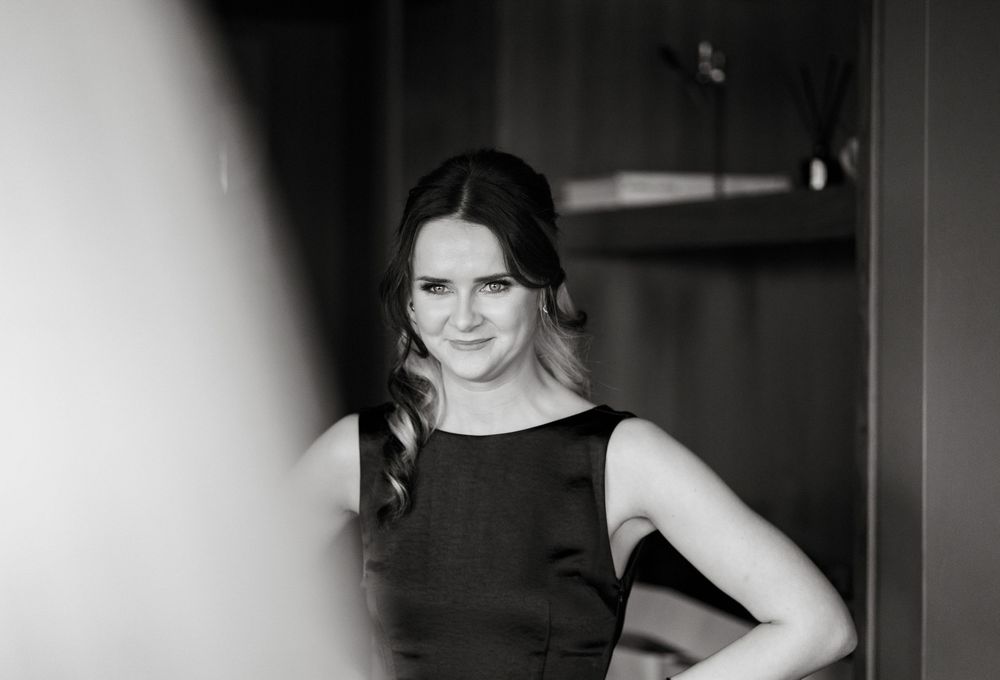 Black and white portrait of a smiling woman in a sleeveless dark dress, posing elegantly indoors.