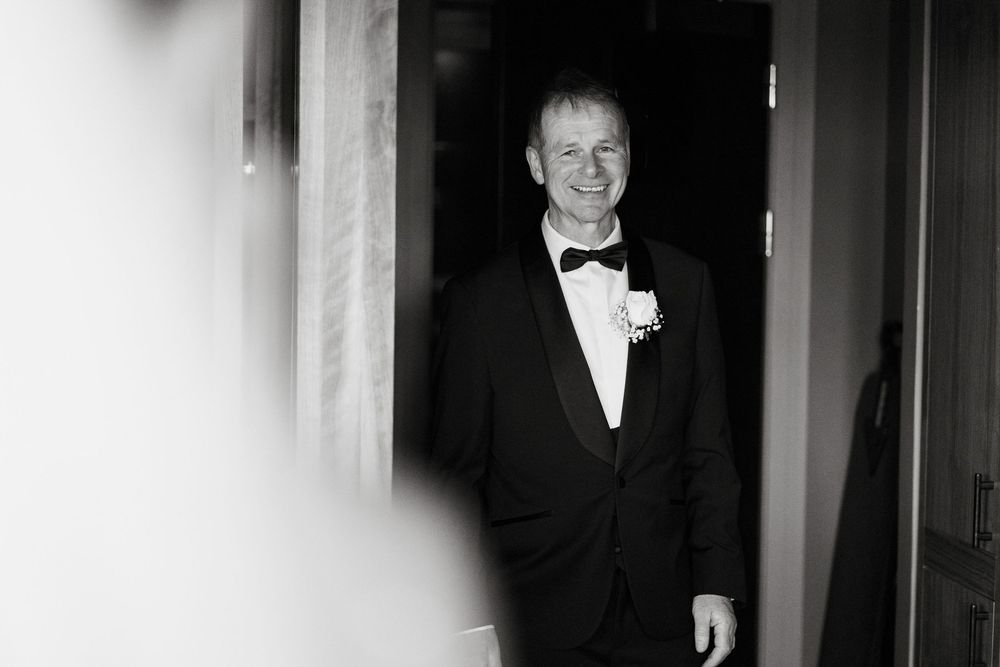 Groom in black tuxedo and bow tie smiling in doorway, black and white wedding photography.