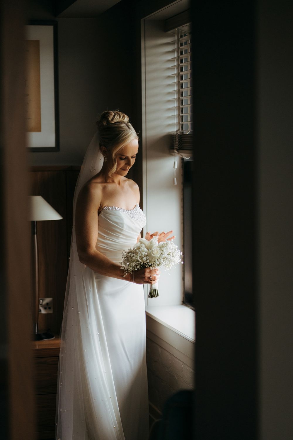 Bride in white strapless gown holding baby's breath bouquet, standing by window in soft natural light.