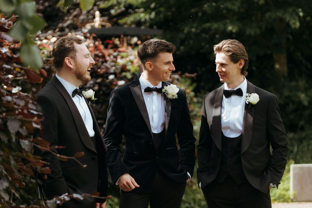 Three groomsmen in black tuxedos with bow ties and white boutonnieres conversing outdoors at a wedding.