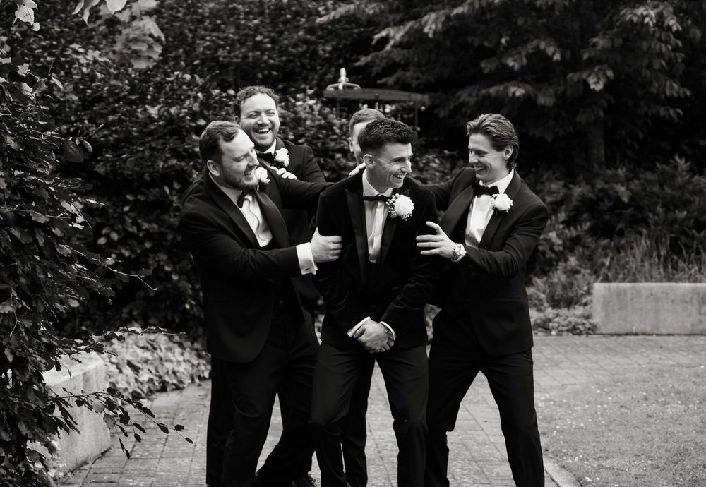 Groomsmen in black tuxedos laughing together outdoors at a wedding celebration.