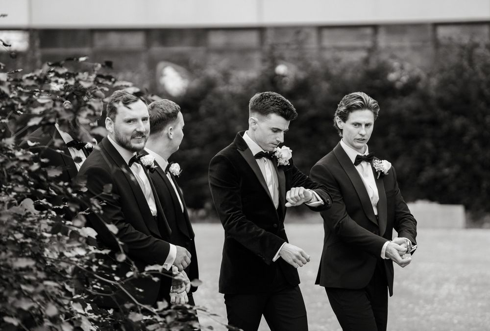 Groomsmen in black tuxedos checking watches outdoors before a wedding ceremony.