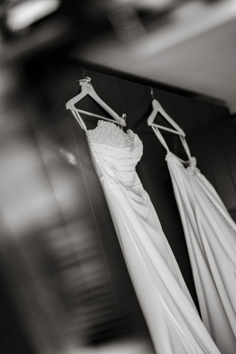 Black and white photo of two elegant wedding dresses hanging on wooden hangers in a bridal boutique.