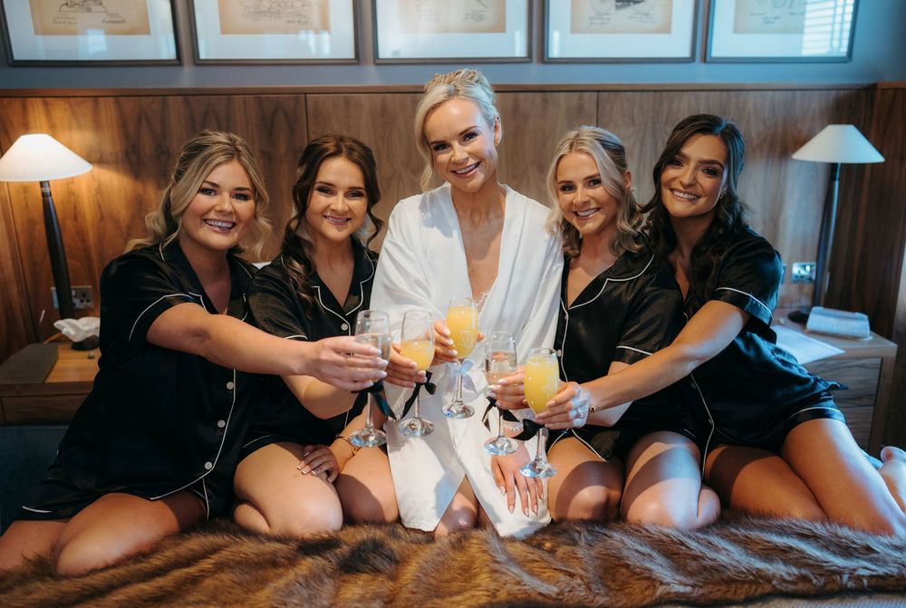 Bride in white robe toasting with four bridesmaids in black satin pajamas on a bed before the wedding.