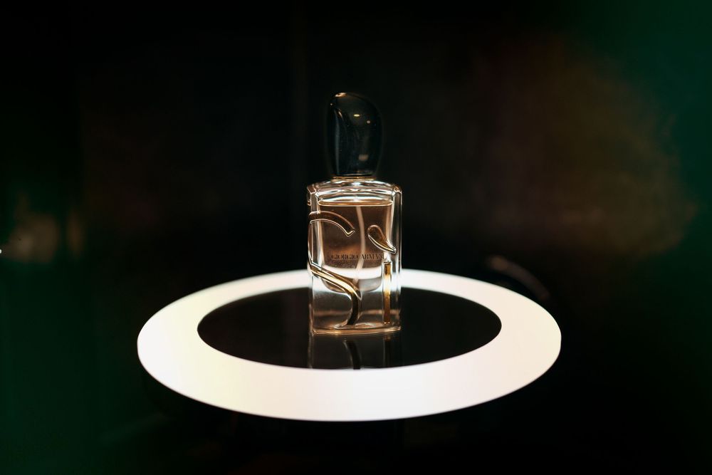 A clear glass perfume bottle displayed on a illuminated circular platform against a dark green background.