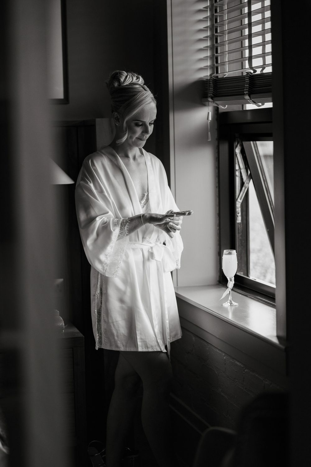 A woman in a white robe checks her phone by a window with a champagne glass nearby.