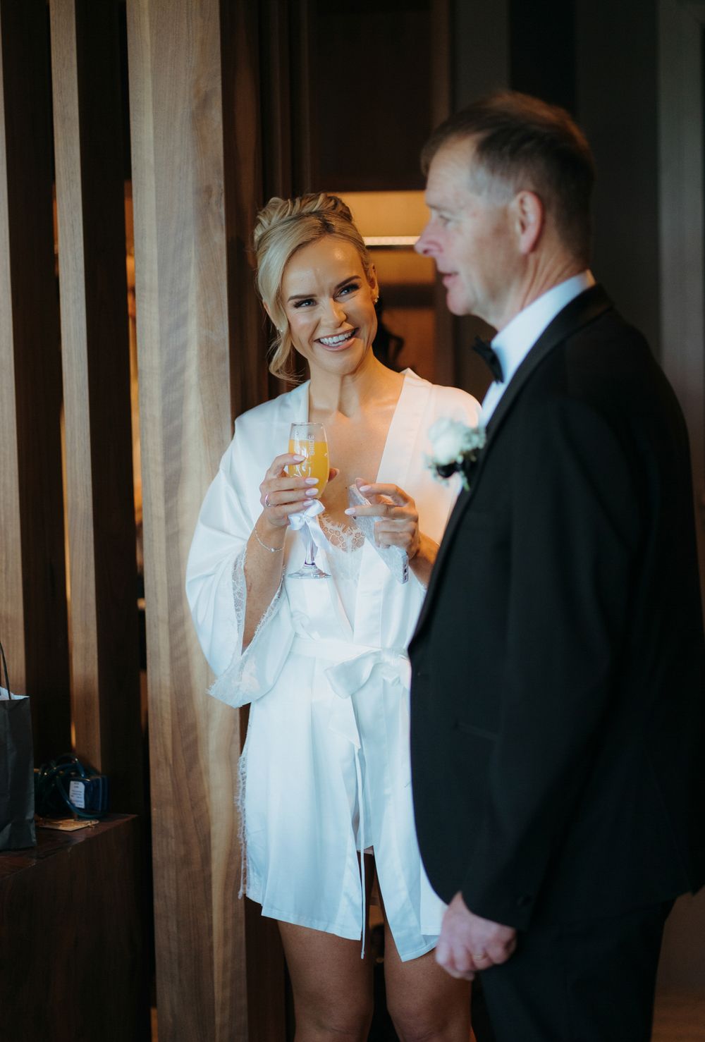 Bride in white satin robe holding champagne flute smiles at groom in black tuxedo during wedding morning preparations.