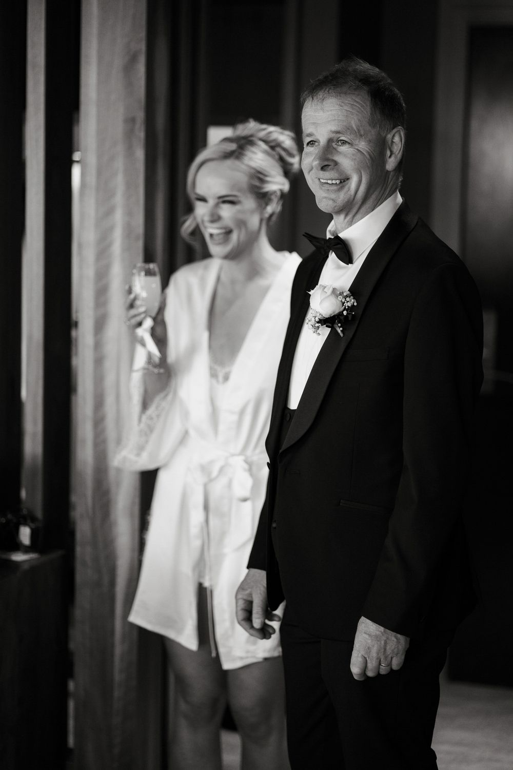 Black and white photo of a smiling bride in a white robe with her father in a tuxedo on wedding day.
