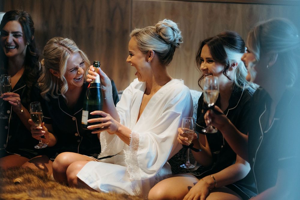 Bride in white robe laughing with bridesmaids holding champagne glasses during a fun bridal party getting ready celebration.