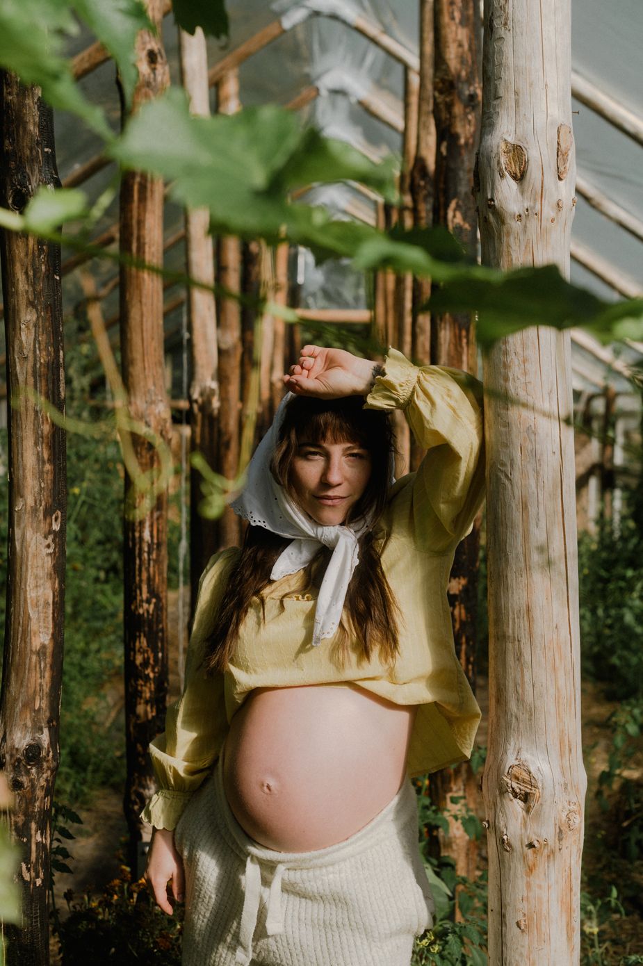 Pregnant woman in yellow crop top poses among rustic wooden poles in a sunlit greenhouse setting.