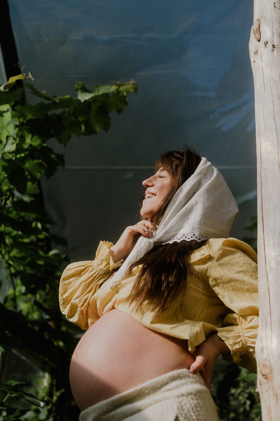 Smiling pregnant woman in yellow dress and white headscarf enjoying sunlight outdoors near green foliage.