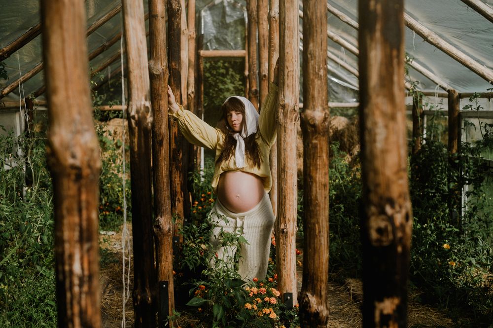 Pregnant woman in hooded outfit standing among wooden poles in a lush greenhouse.