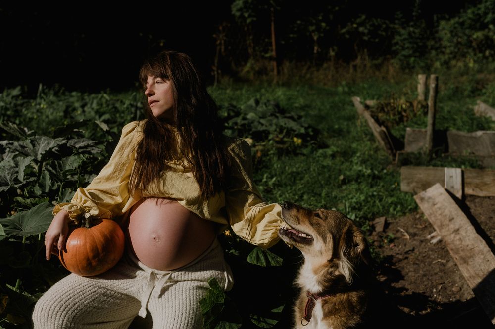 Pregnant woman sitting outdoors with a pumpkin and a cat beside her.