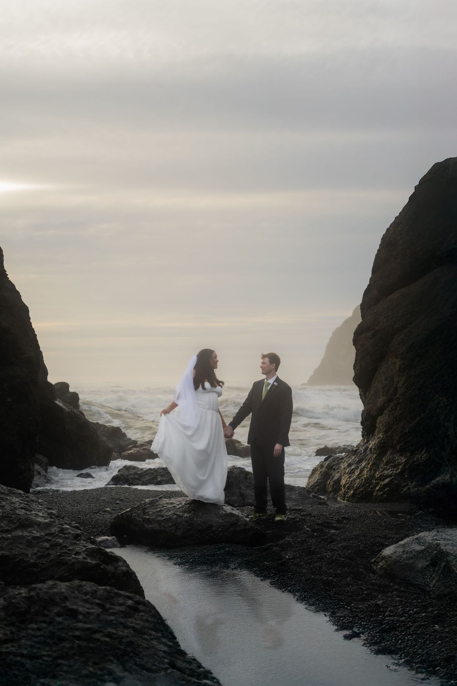 Bride and groom hold hands on rocky ocean shore at sunset, waves crashing behind them in wedding attire.