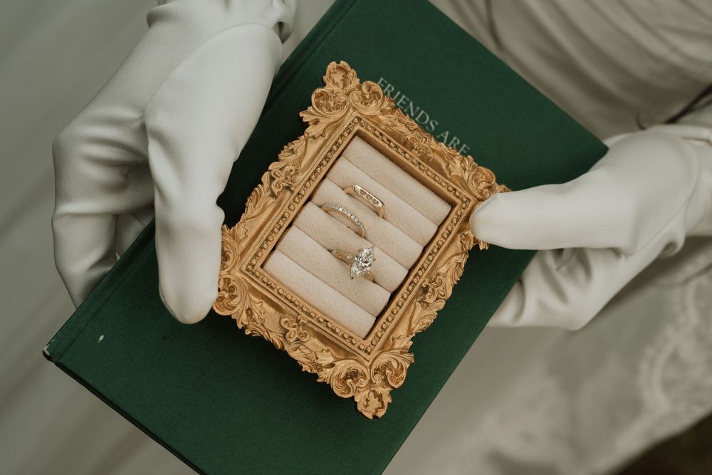 Gloved hands holding a green book with a gold ornate frame displaying elegant diamond wedding rings.