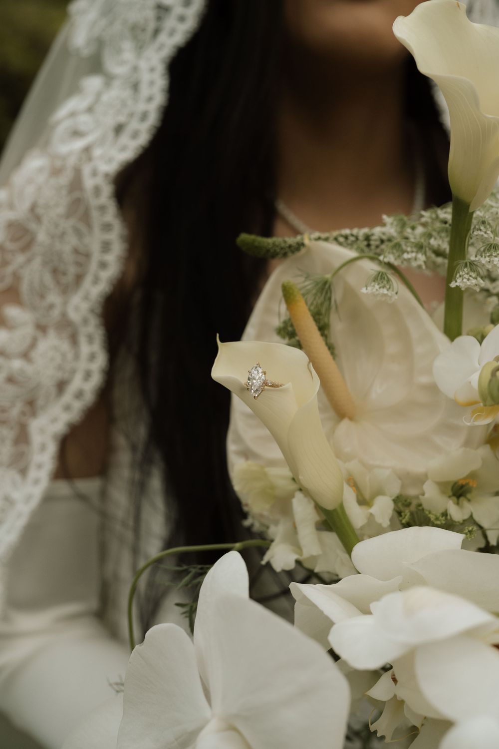 Bride holding white calla lily bouquet with diamond engagement ring displayed among elegant white flowers and lace veil.