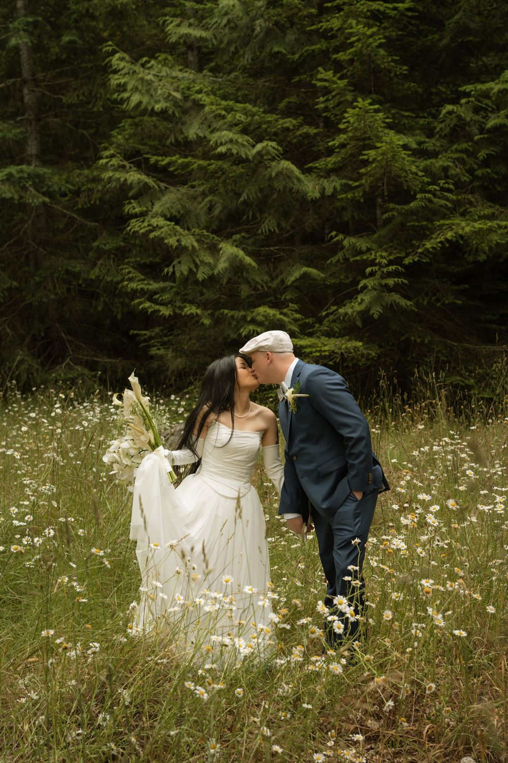 Bride and groom share a kiss in a wildflower meadow surrounded by tall evergreen trees.