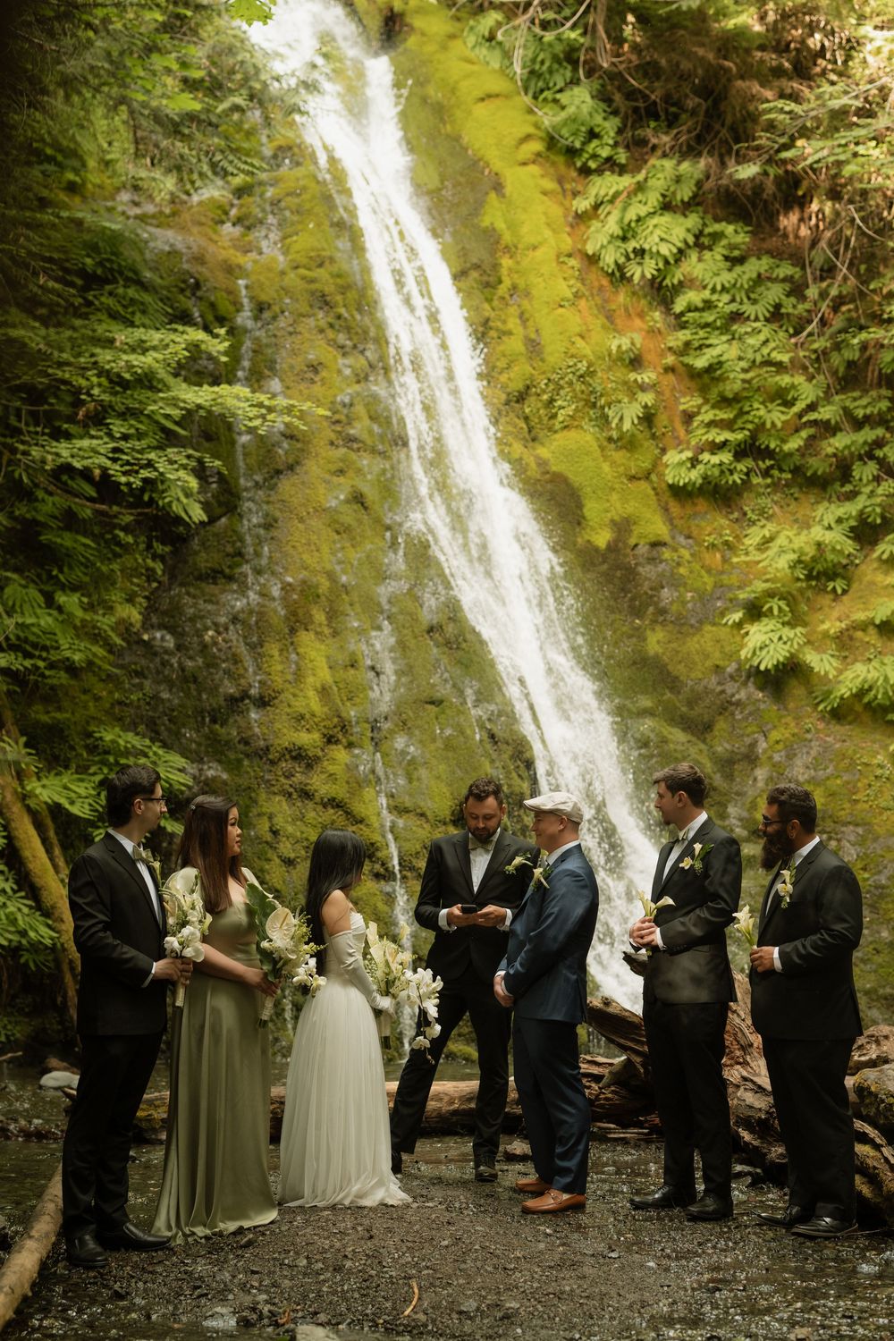 Wedding party stands before a cascading waterfall in a lush forest during an intimate elopement ceremony.