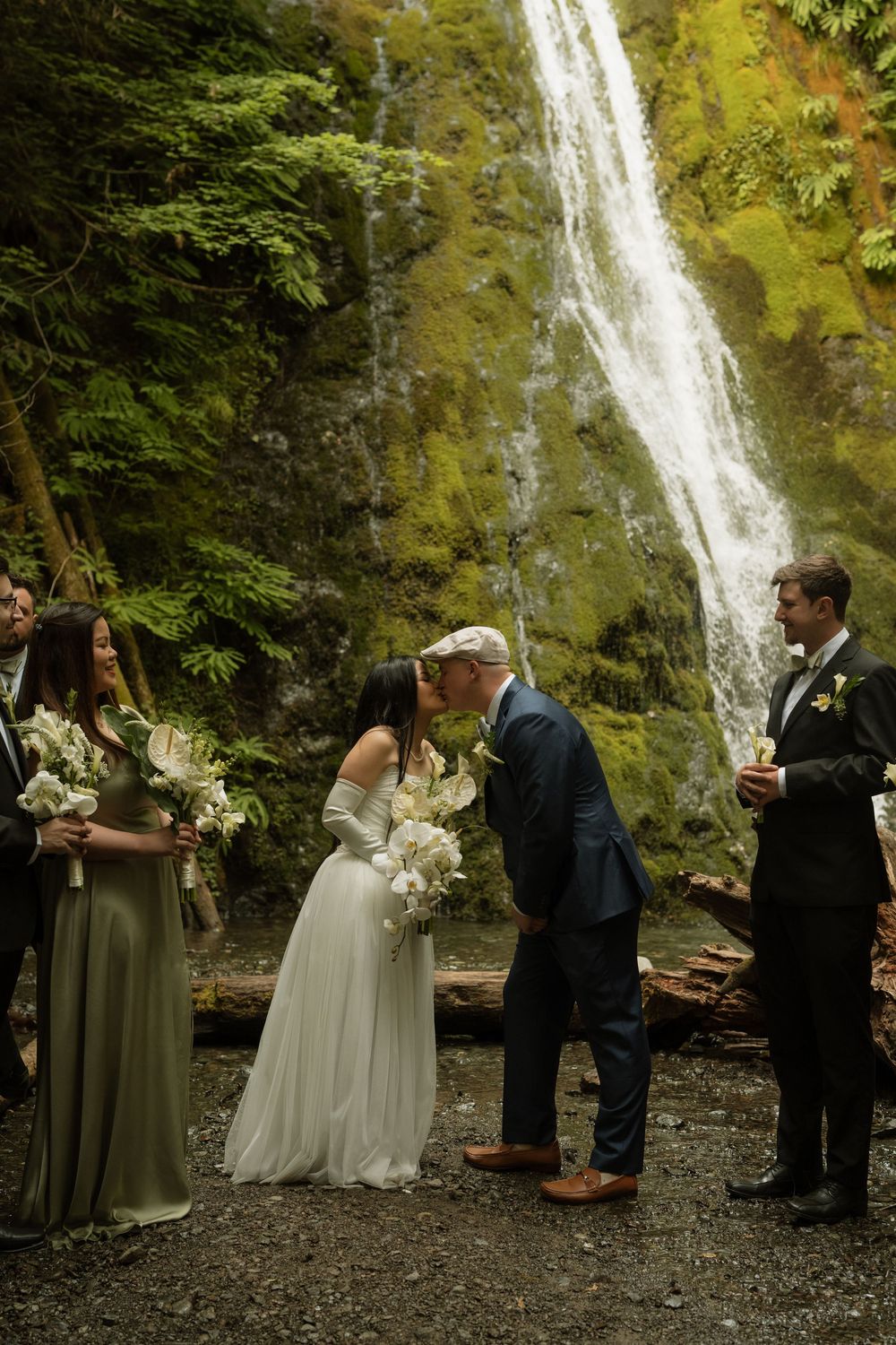 Bride and groom share first kiss at outdoor waterfall wedding ceremony with wedding party beside them.
