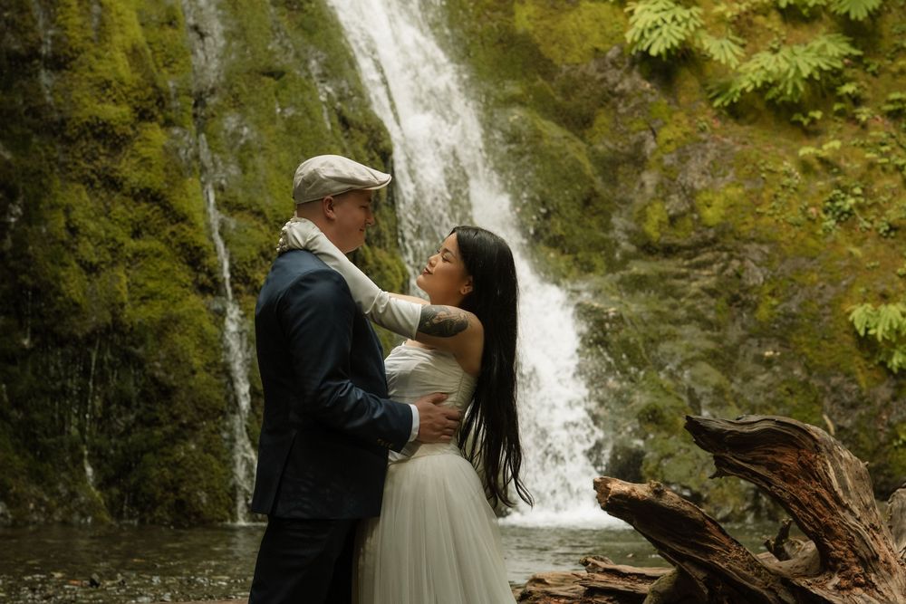 Couple embracing romantically in front of a lush mossy waterfall, bride in white gown and groom in navy suit.