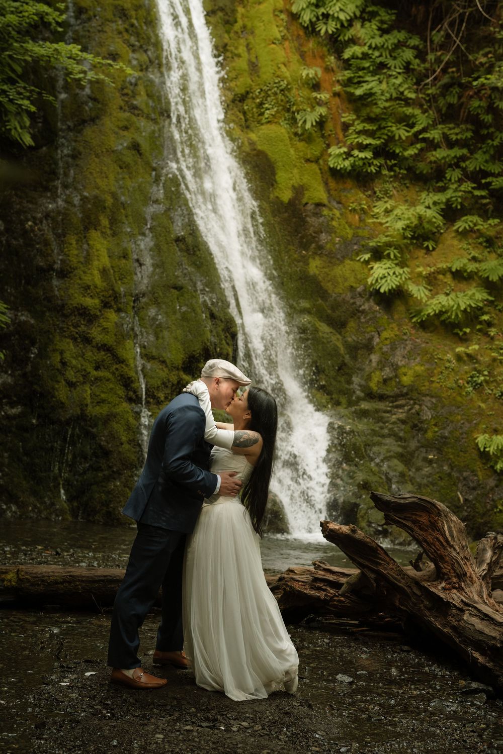 Bride and groom share romantic kiss in front of stunning mossy waterfall in lush forest setting.