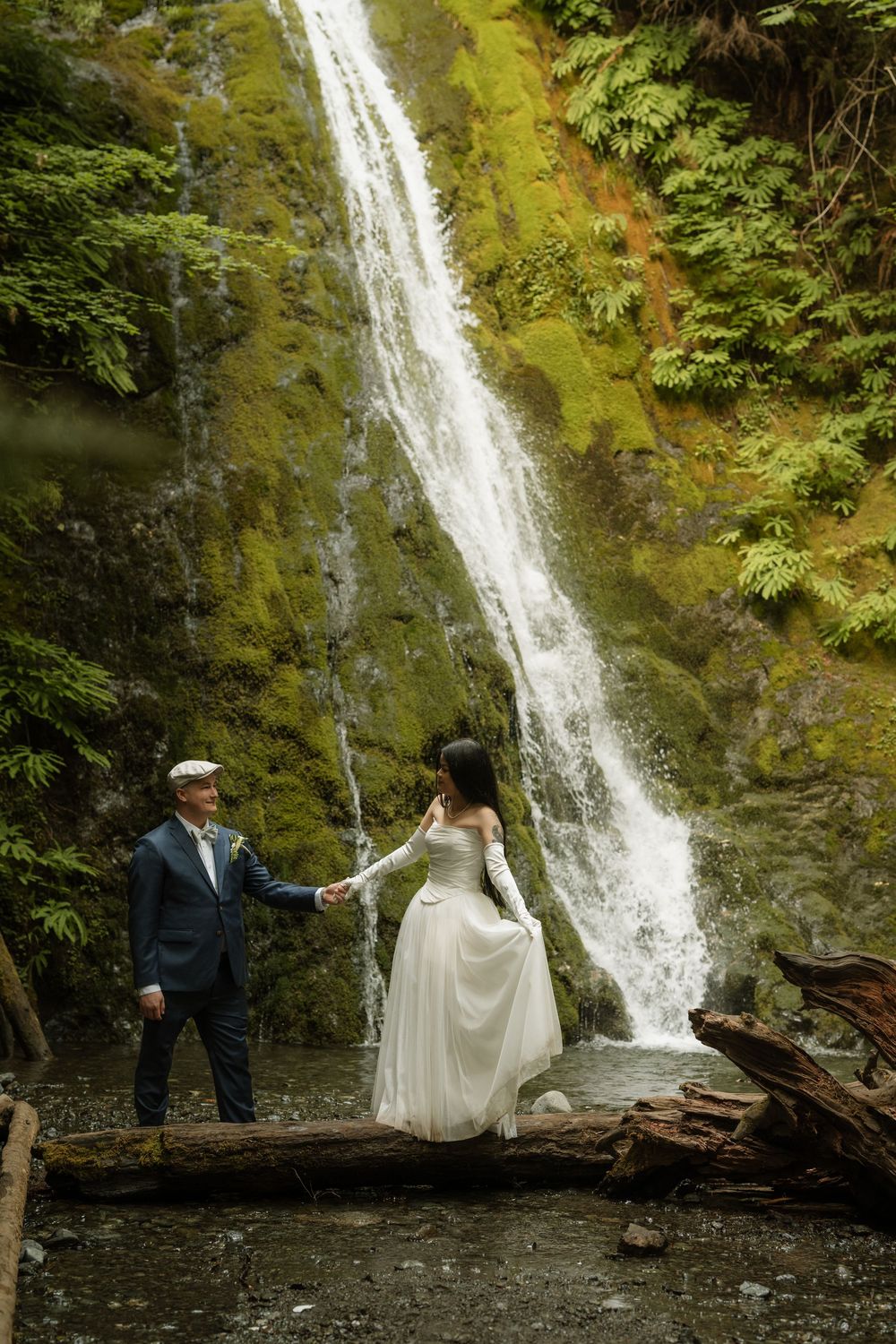 Bride and groom hold hands on a log before a lush waterfall in a green forest during their outdoor wedding.