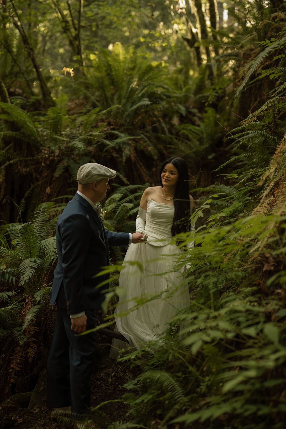 Couple in wedding attire share a tender moment surrounded by ferns in forest.