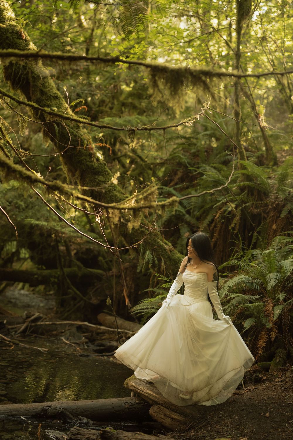 Bride in white ball gown and gloves poses in lush green forest with ferns and mossy trees.